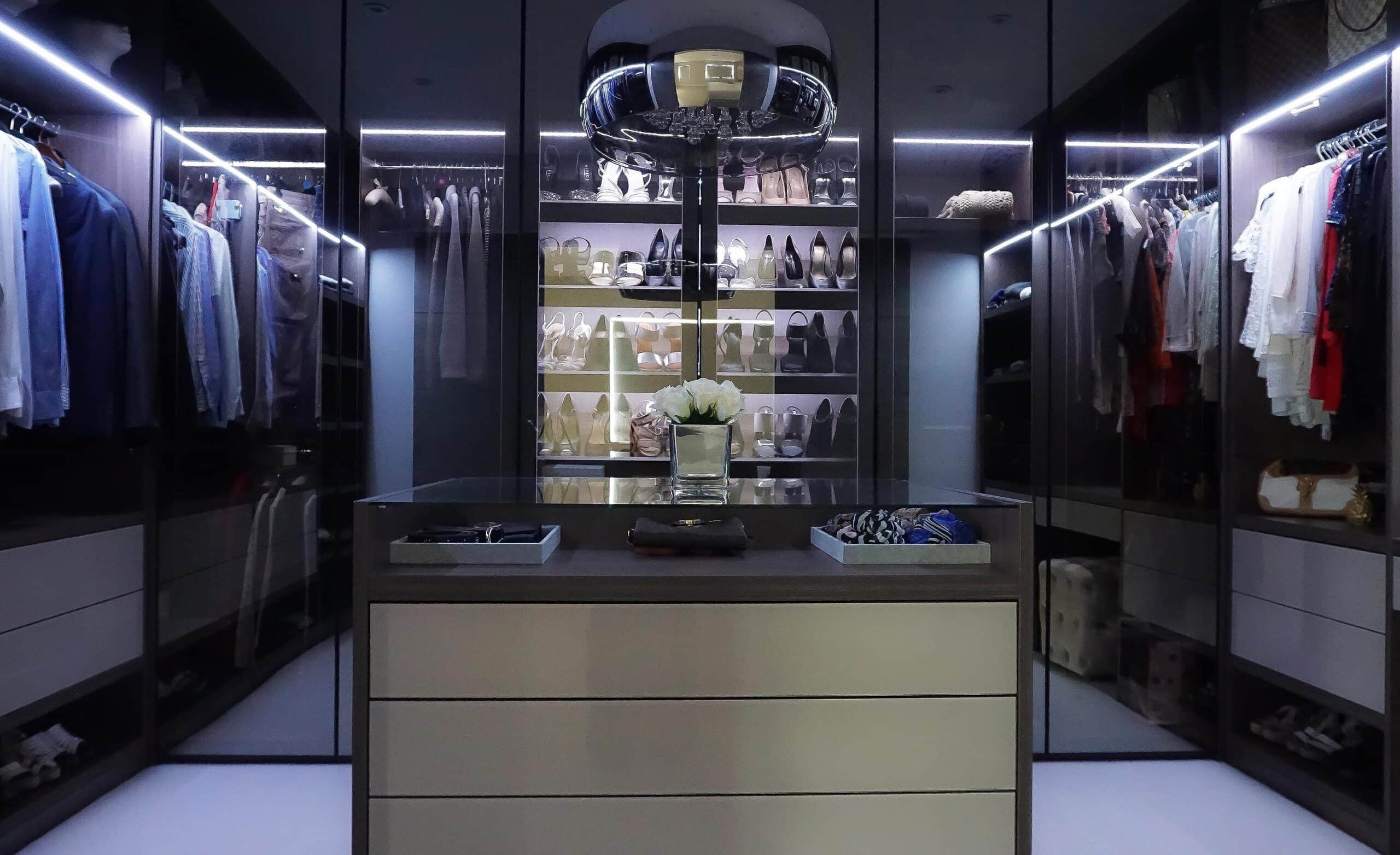 Dark walk-in wardrobe with LED-lit hanging rails, glass doors and a central island