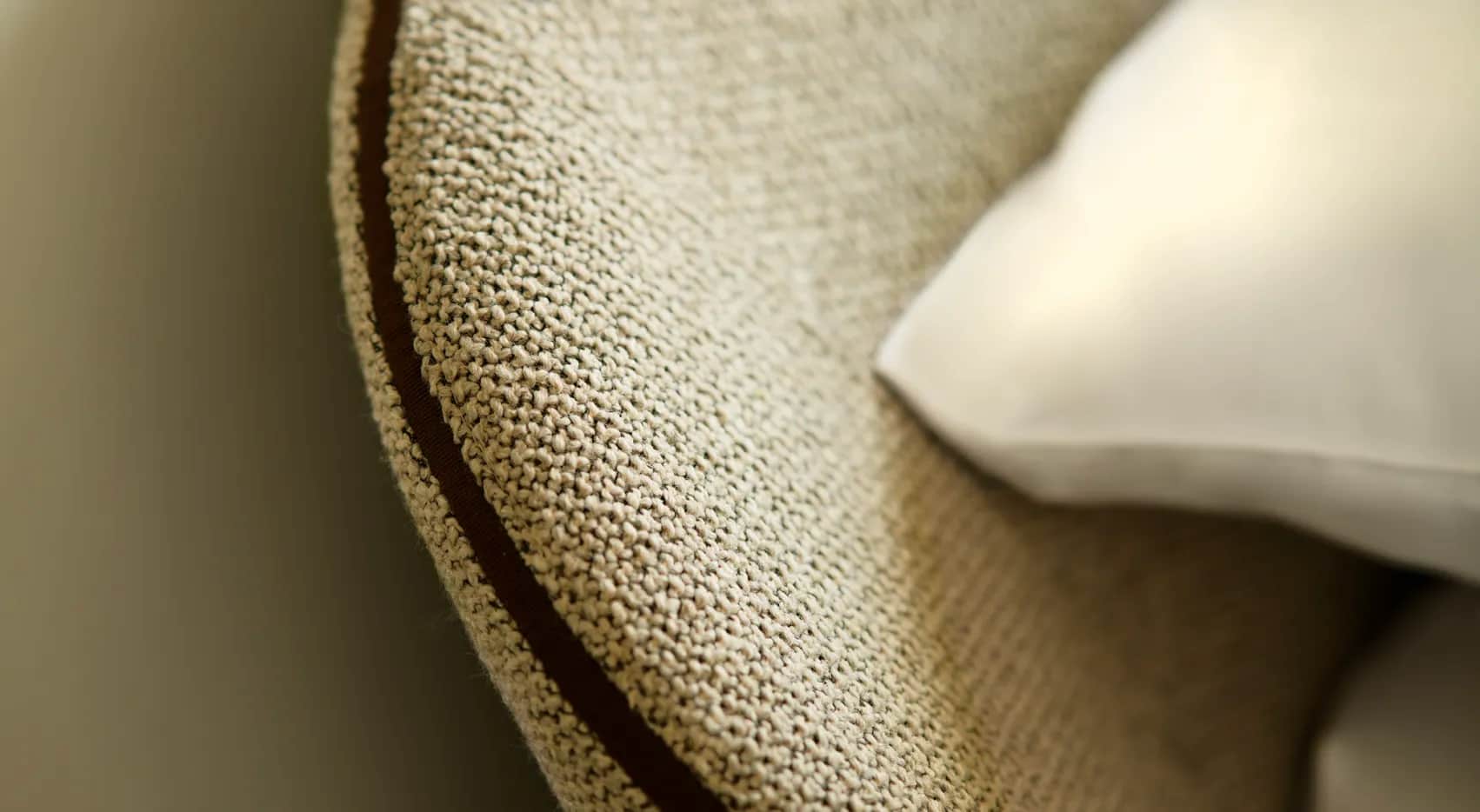 Close-up of cream textured upholstery fabric with brown piping and a white pillow