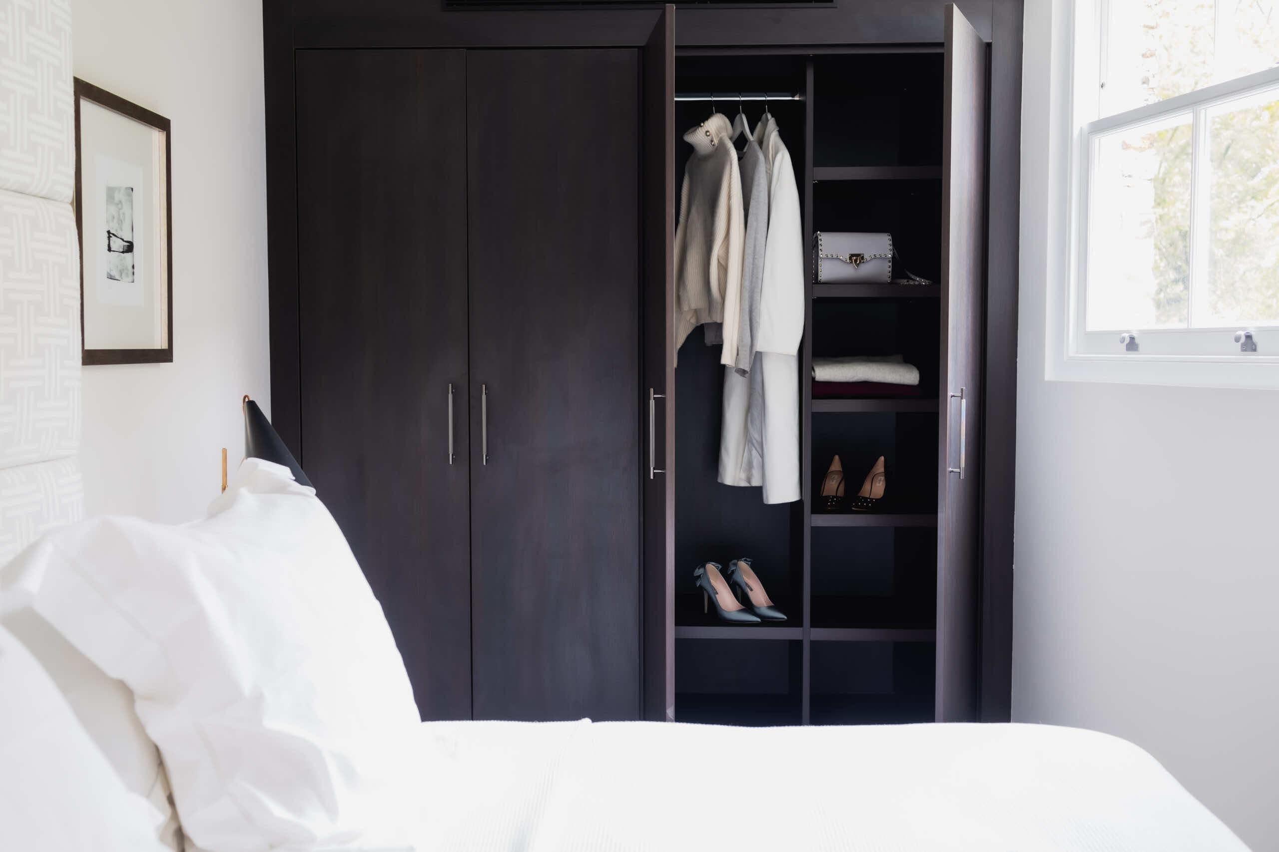Dark wood fitted wardrobe with one door open beside a bed with white linen
