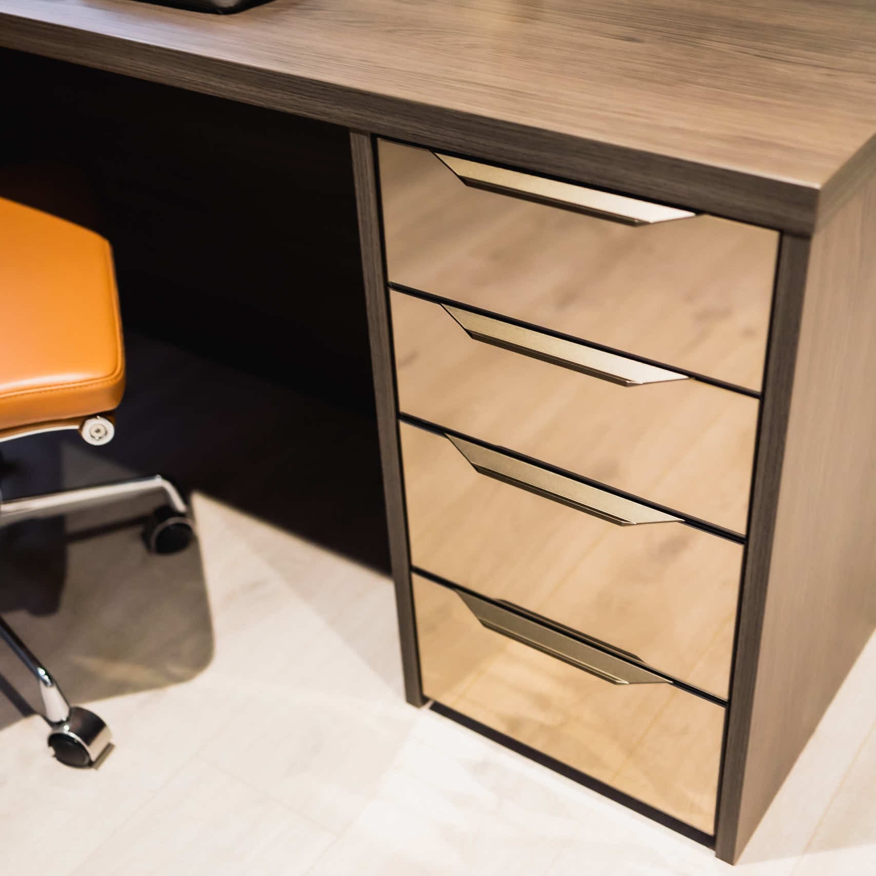 Study desk with mirrored drawer fronts and brushed metal handles beside an orange chair