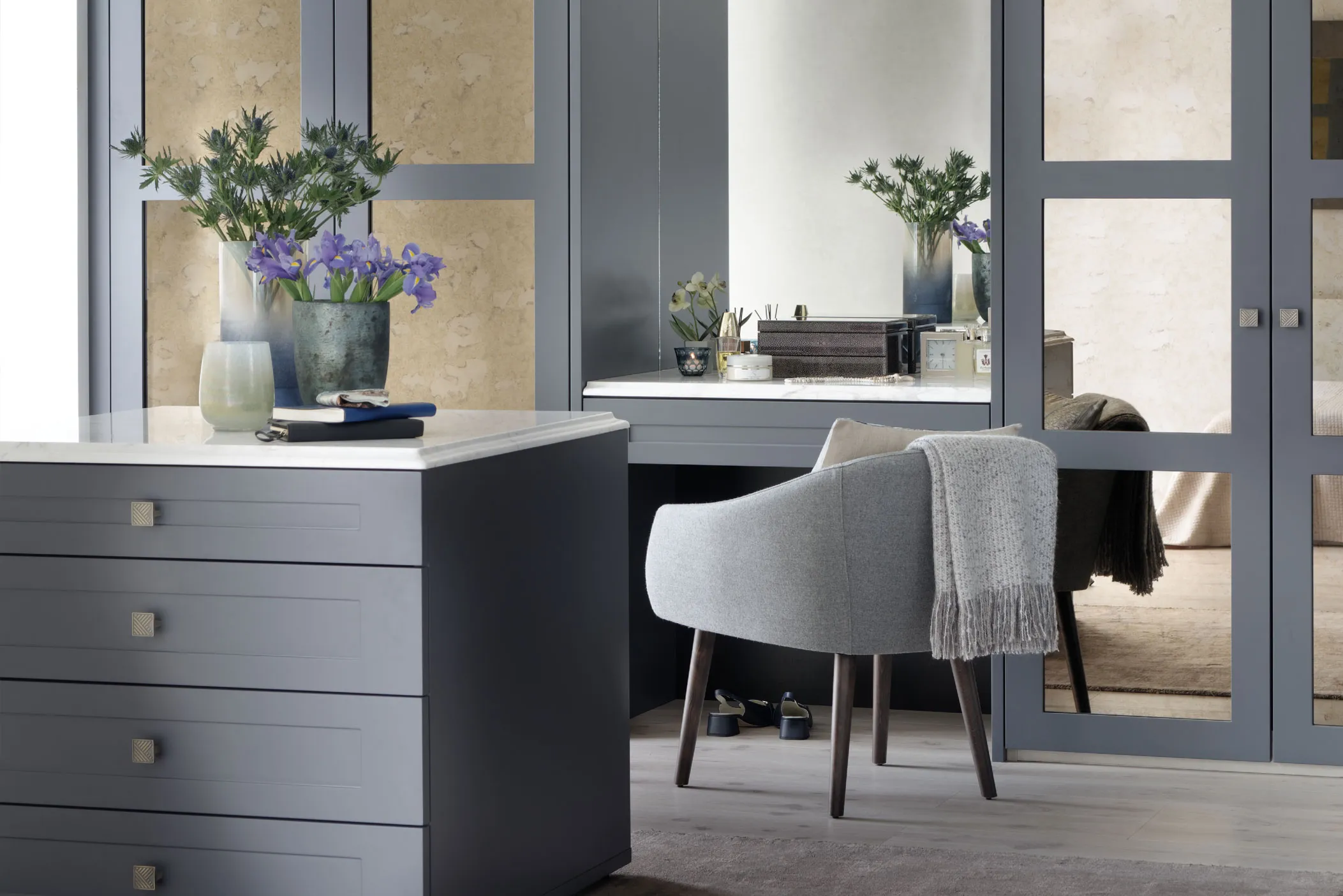 Grey fitted dressing table and drawer unit beside glazed wardrobe doors, with armchair and flowers