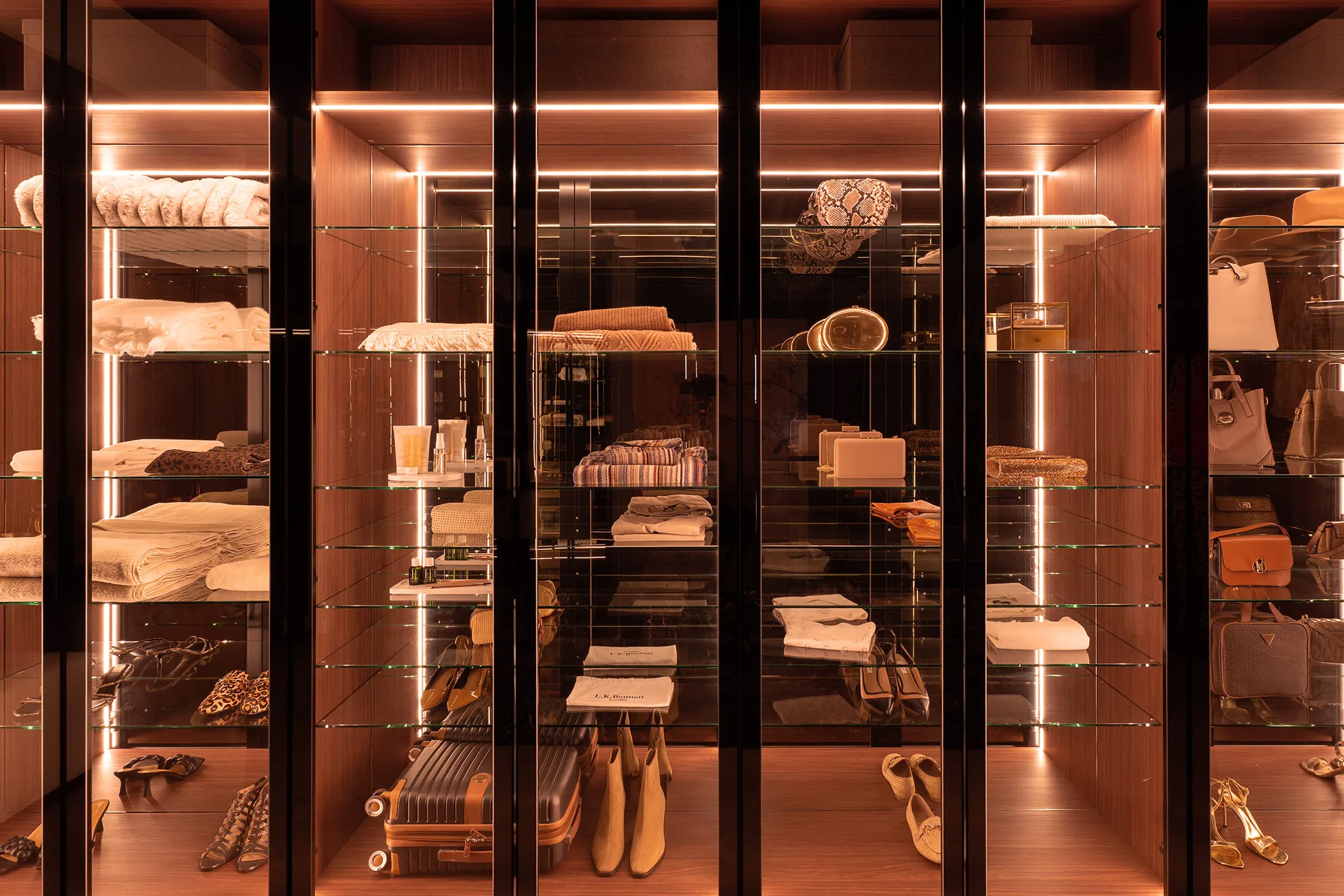 Glass-fronted dressing room cabinets with LED lighting displaying handbags, folded knitwear and luggage