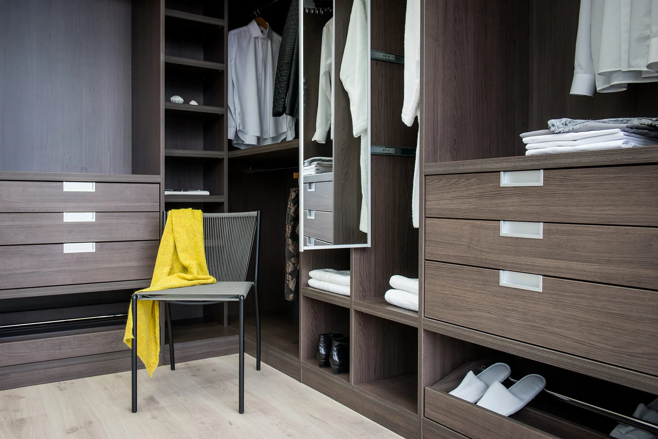 Corner walk-in wardrobe with mirror, drawers, hanging clothes and a yellow throw on a chair