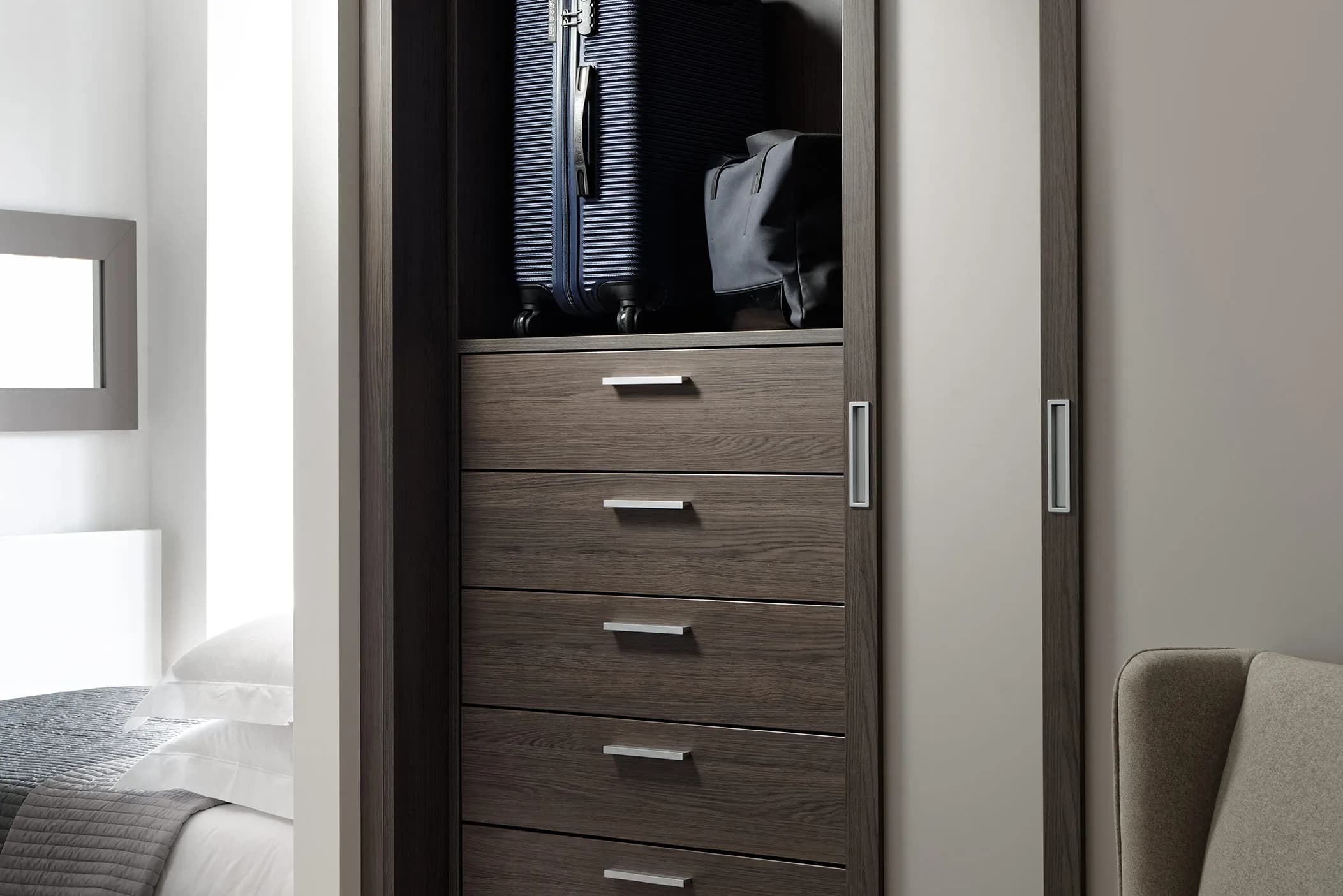 Fitted wardrobe with chrome-handled drawers and a suitcase above, beside a bed