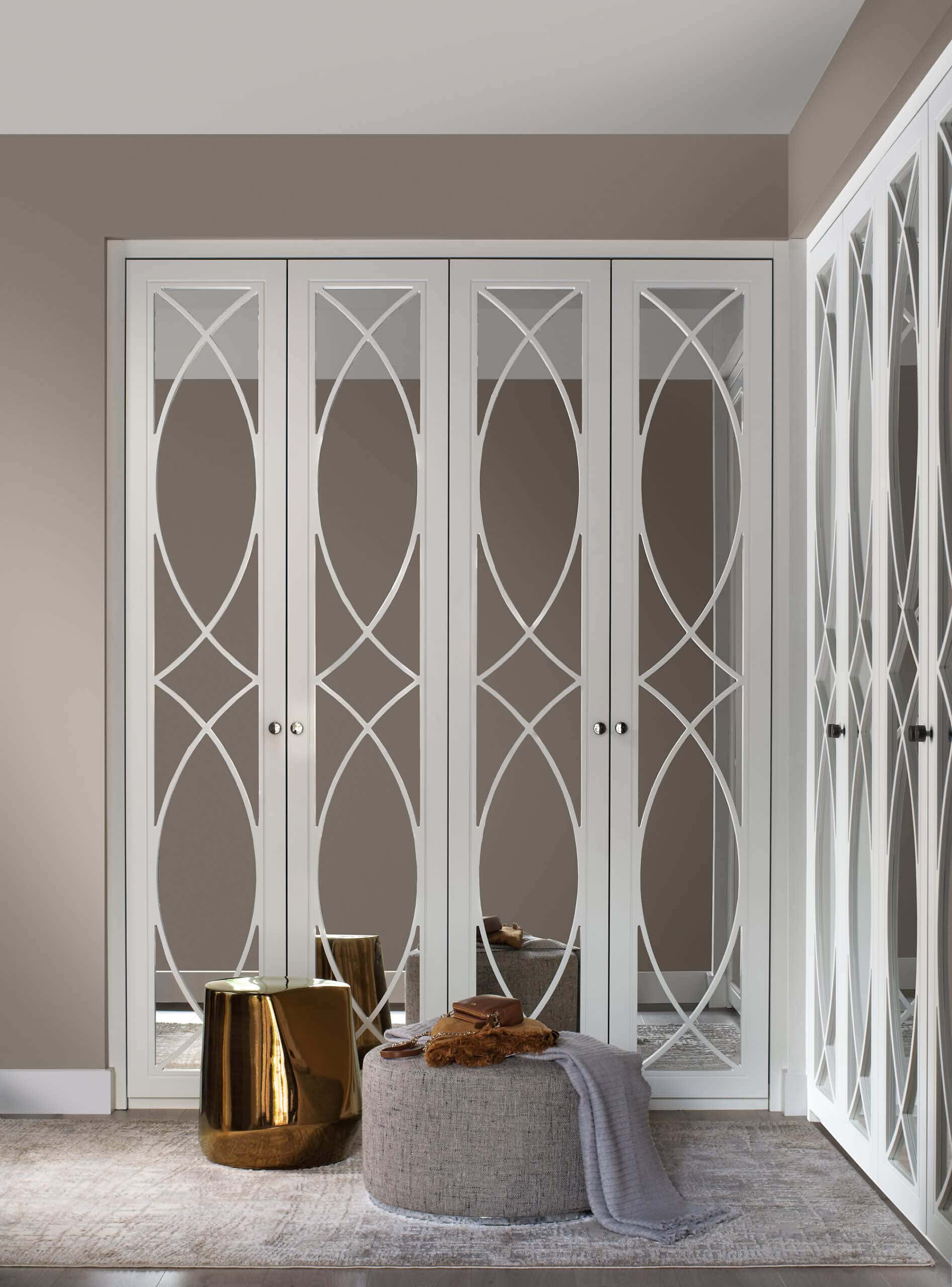 White wardrobe doors with curved mirrored fretwork panels, beside a bronze side table and pouffe