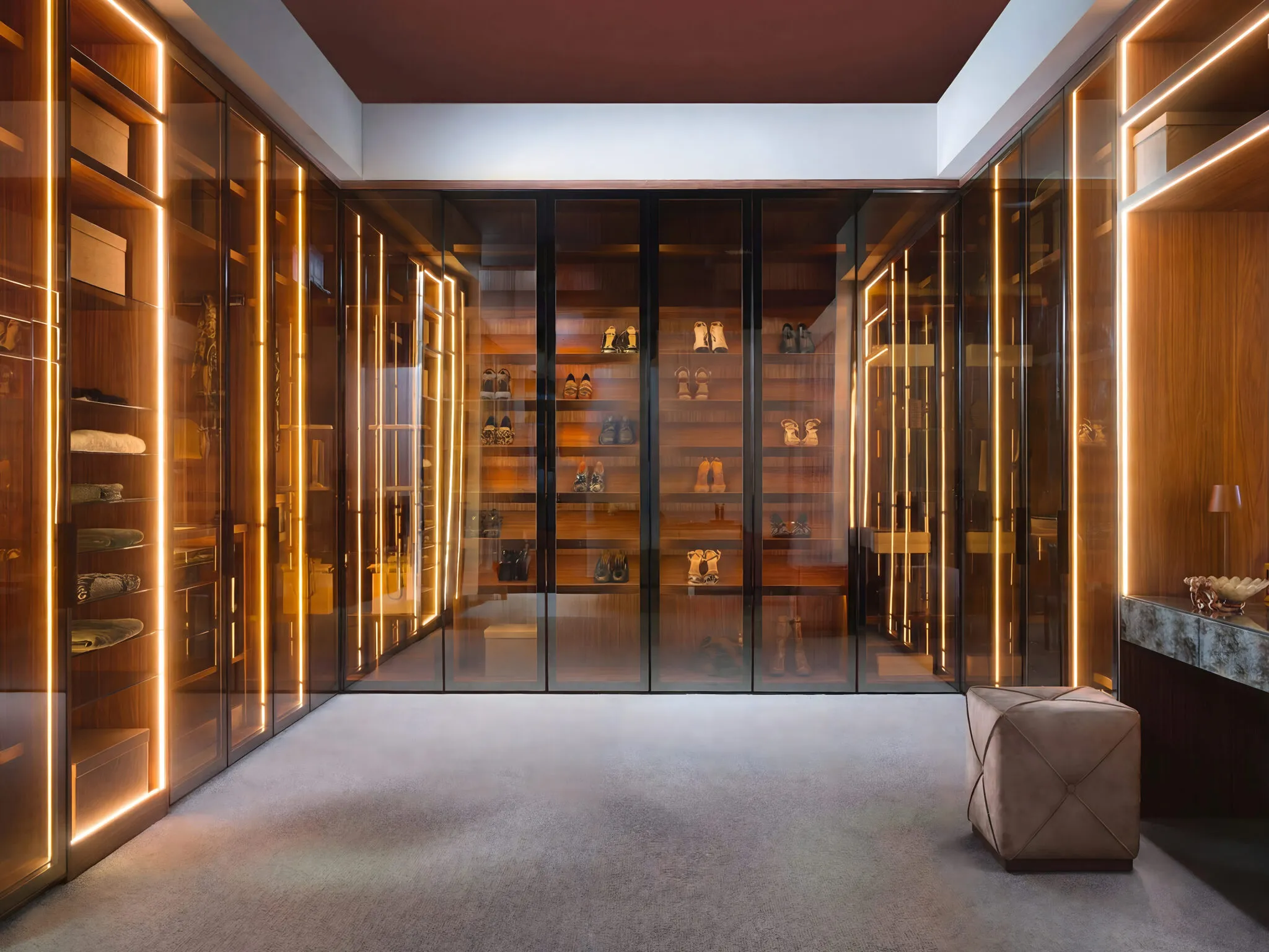 Walk-in wardrobe lined with glass doors, vertical LED strips and a soft pouffe