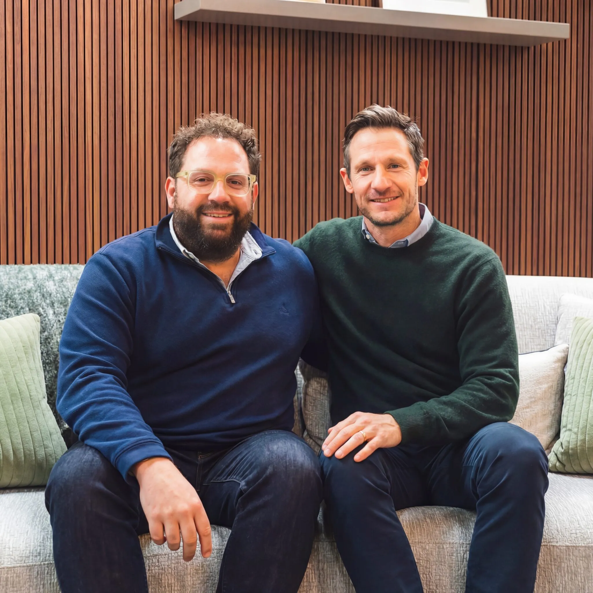Two smiling men sitting together on a sofa in front of a slatted wood wall