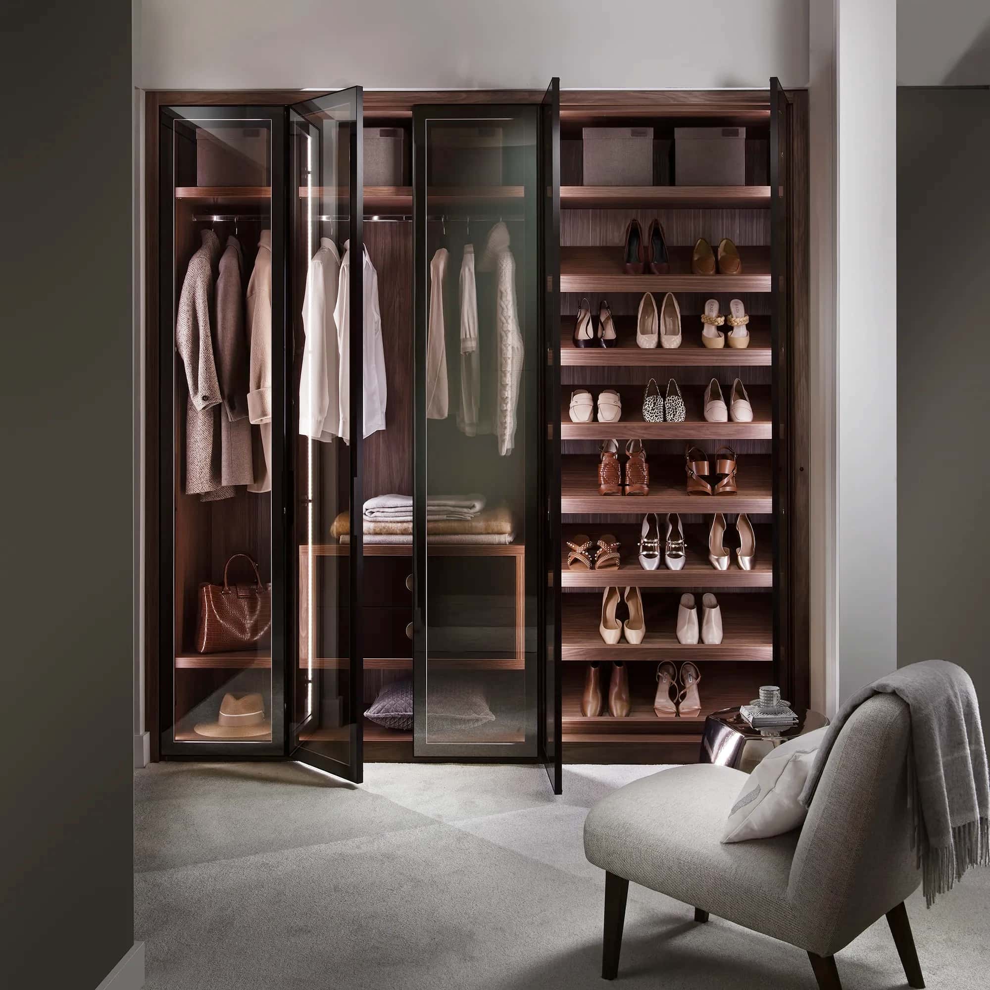 Walk-in wardrobe with smoked glass doors, shoe shelving and hanging rails beside a chair