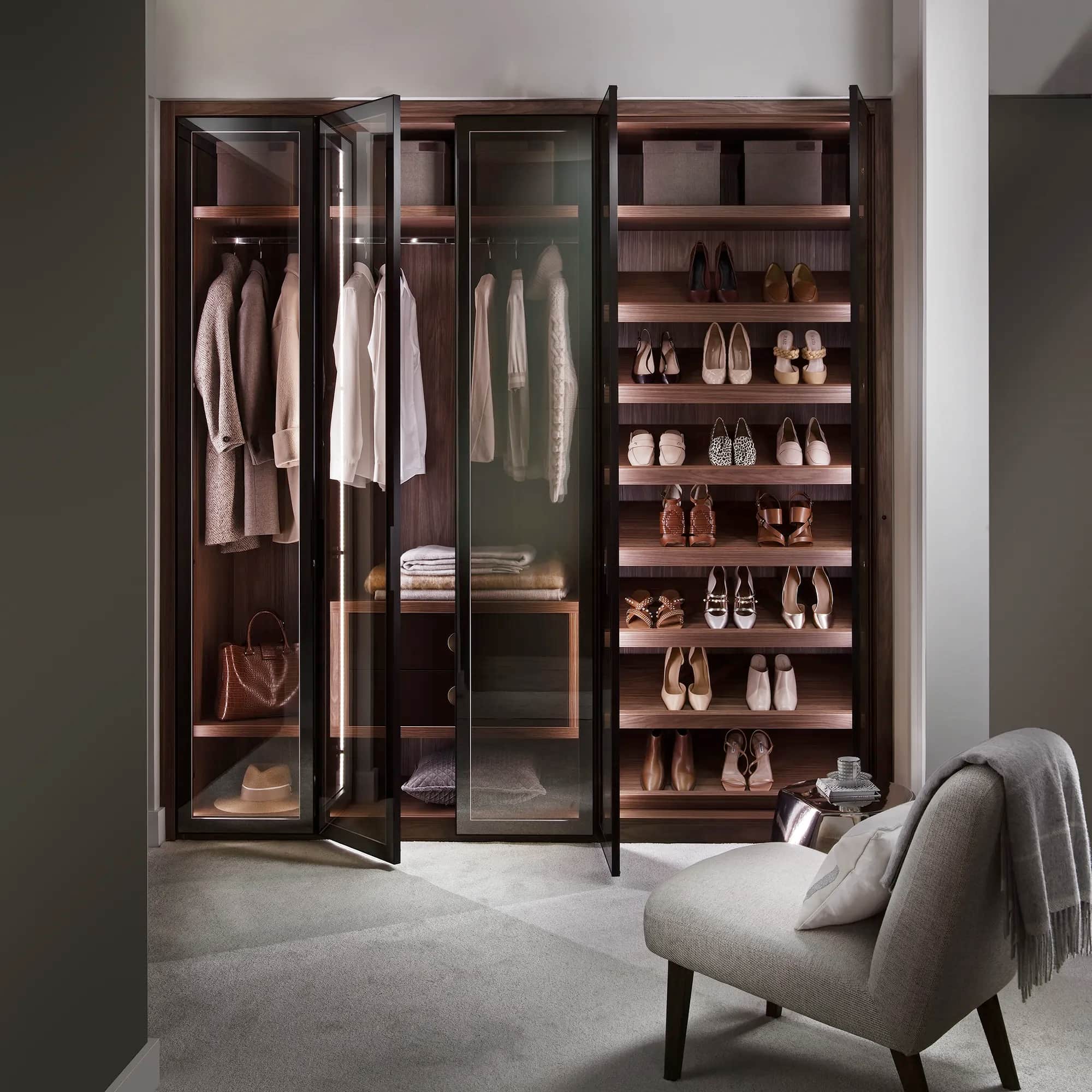 Walk-in wardrobe with smoked glass doors, shoe shelving and hanging rails beside a chair