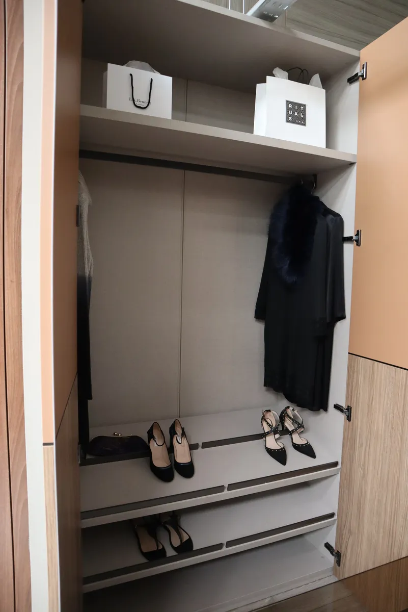 Open wardrobe with shoe drawers, hanging coat and shopping bags behind tan doors