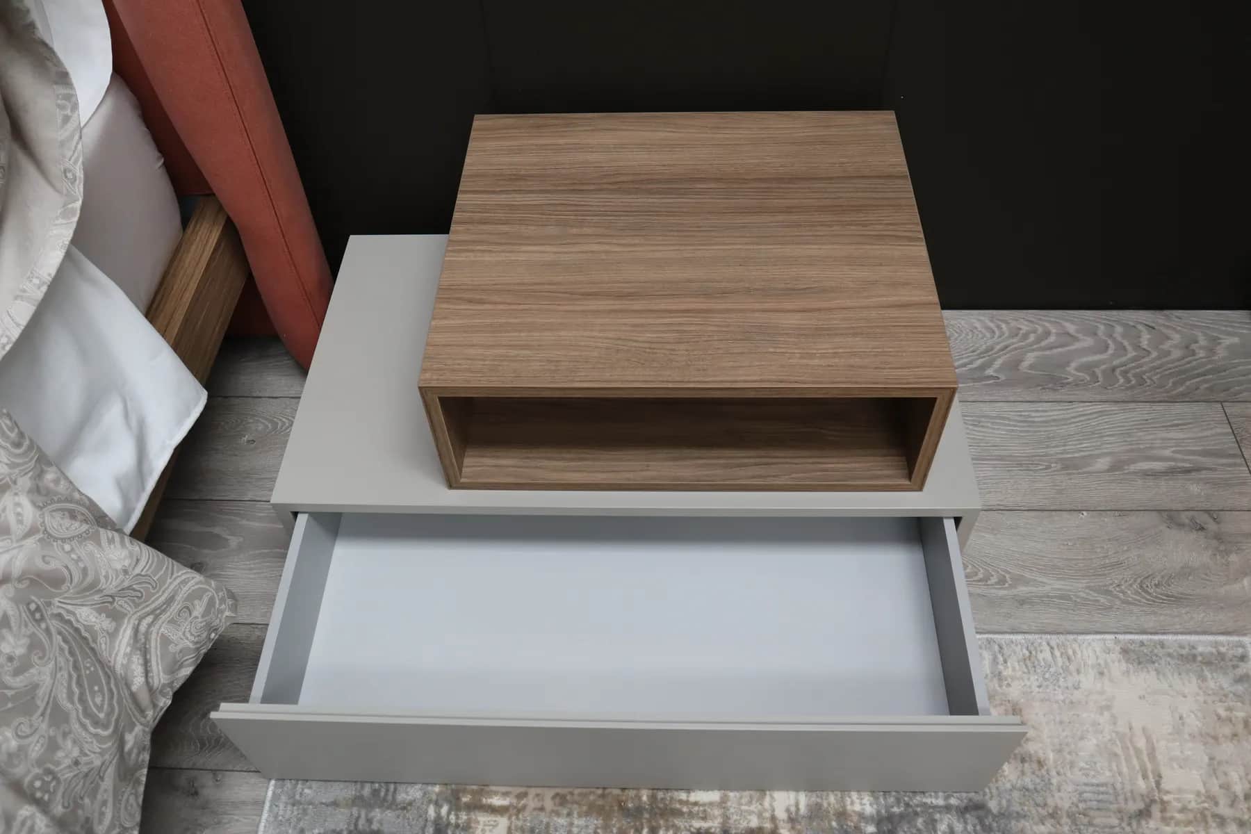 Grey bedside unit with open drawer and walnut open shelf box beside a bed