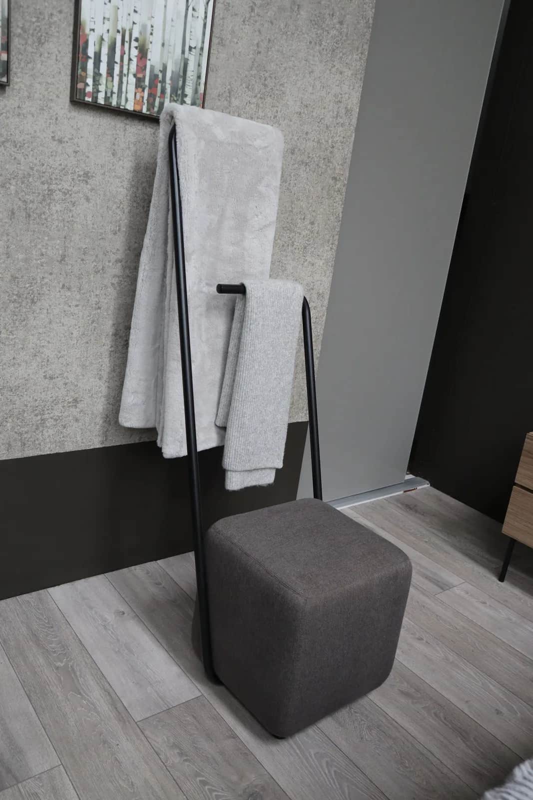 Black towel valet stand with grey towels beside a dark grey upholstered pouffe