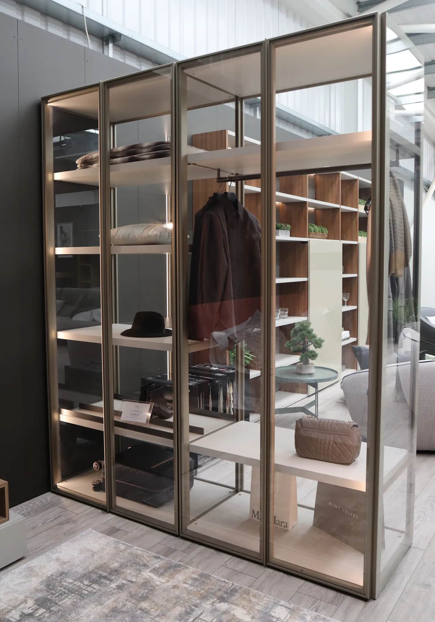 Glass-fronted open wardrobe display with bronze frames, hanging coat, hat and handbag