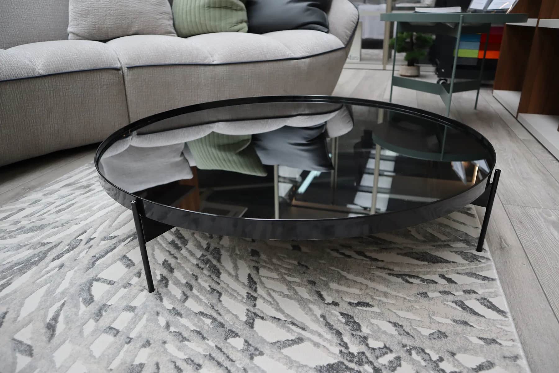 Round dark glass coffee table with black legs on a patterned grey rug beside a sofa