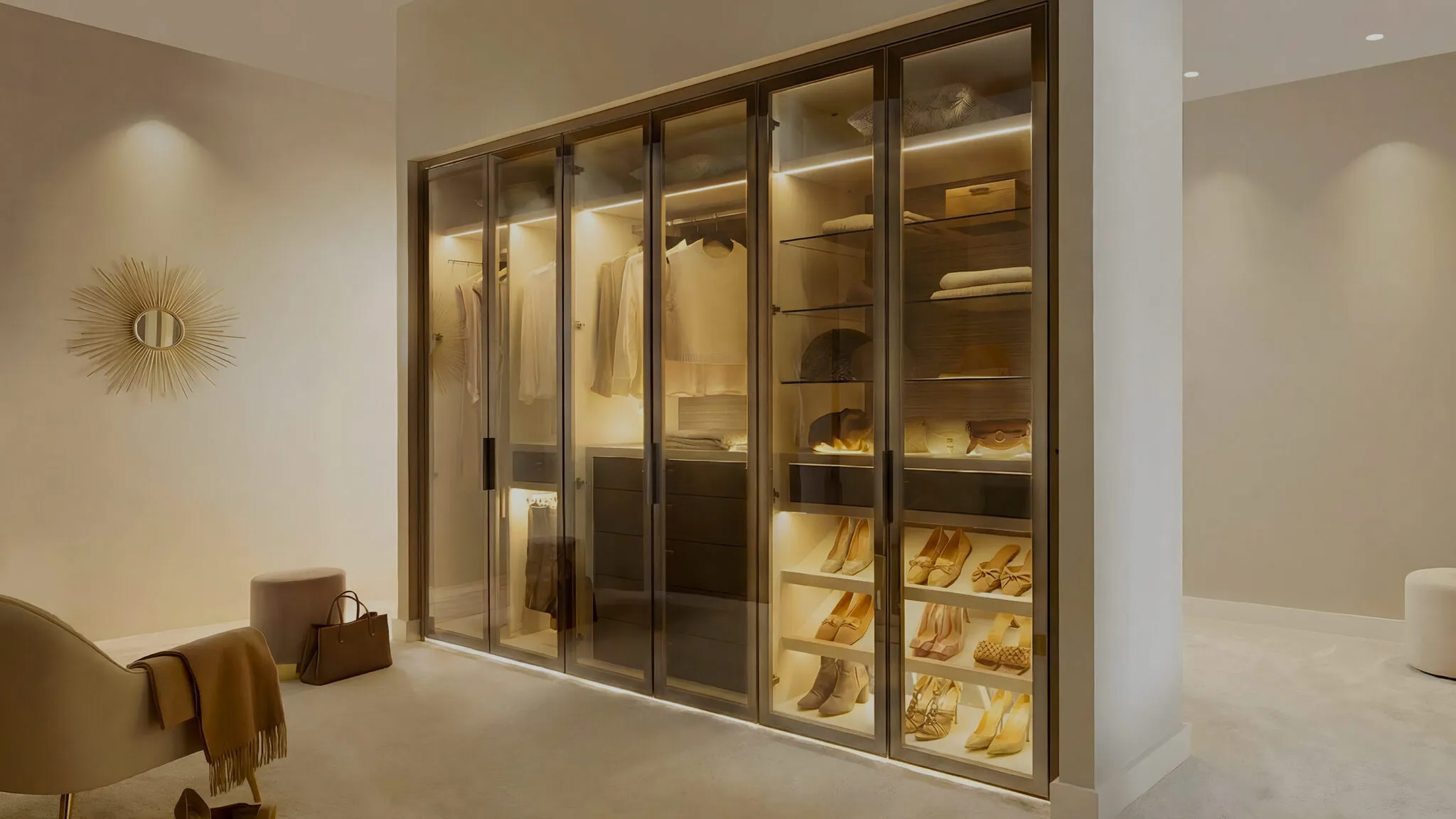 Glass-fronted walk-in wardrobe with LED lighting, shoe shelves and a cream pouffe