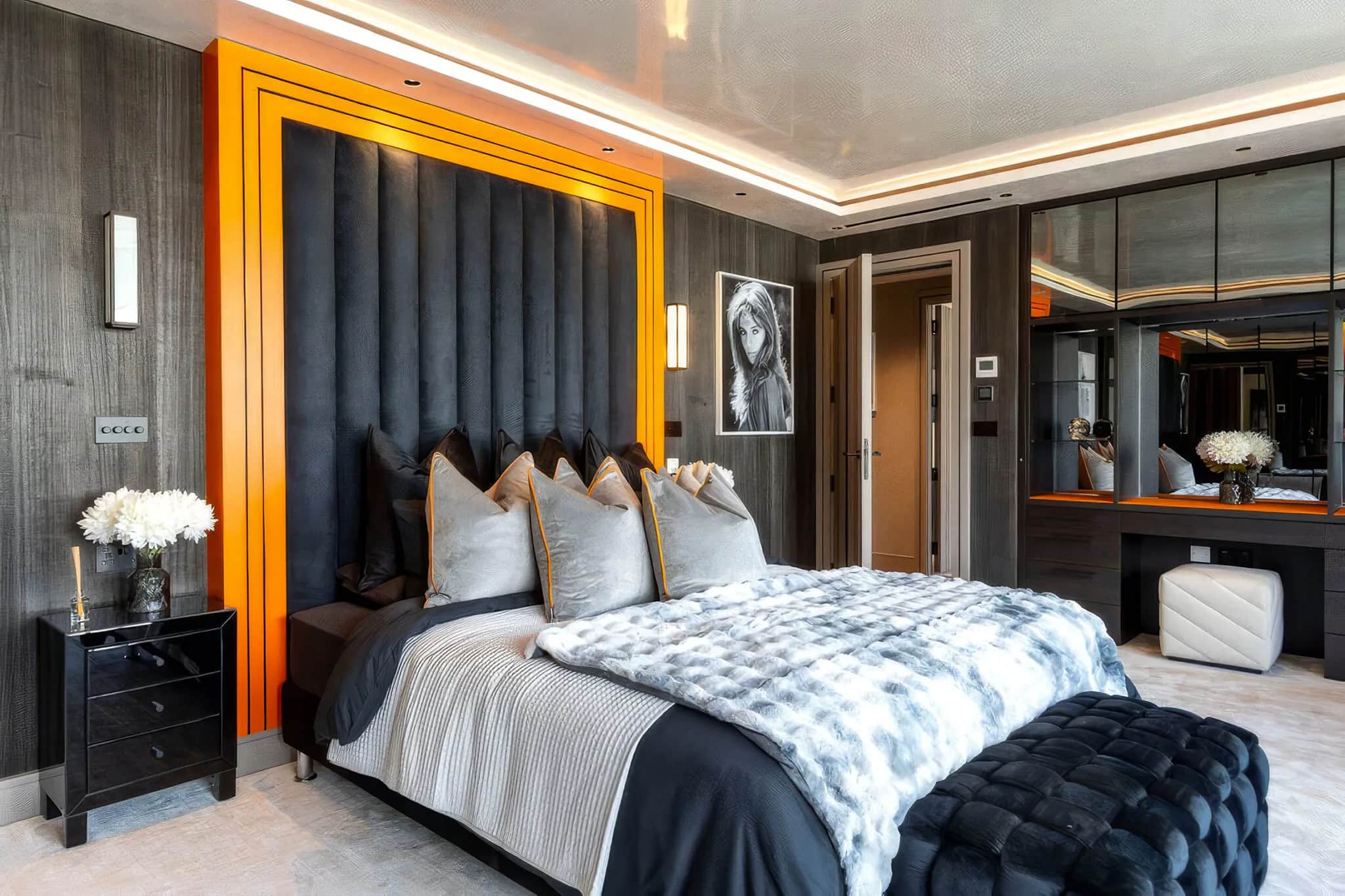 Penthouse bedroom with tall grey padded headboard, orange trim, mirrored wardrobes and a black quilted bench
