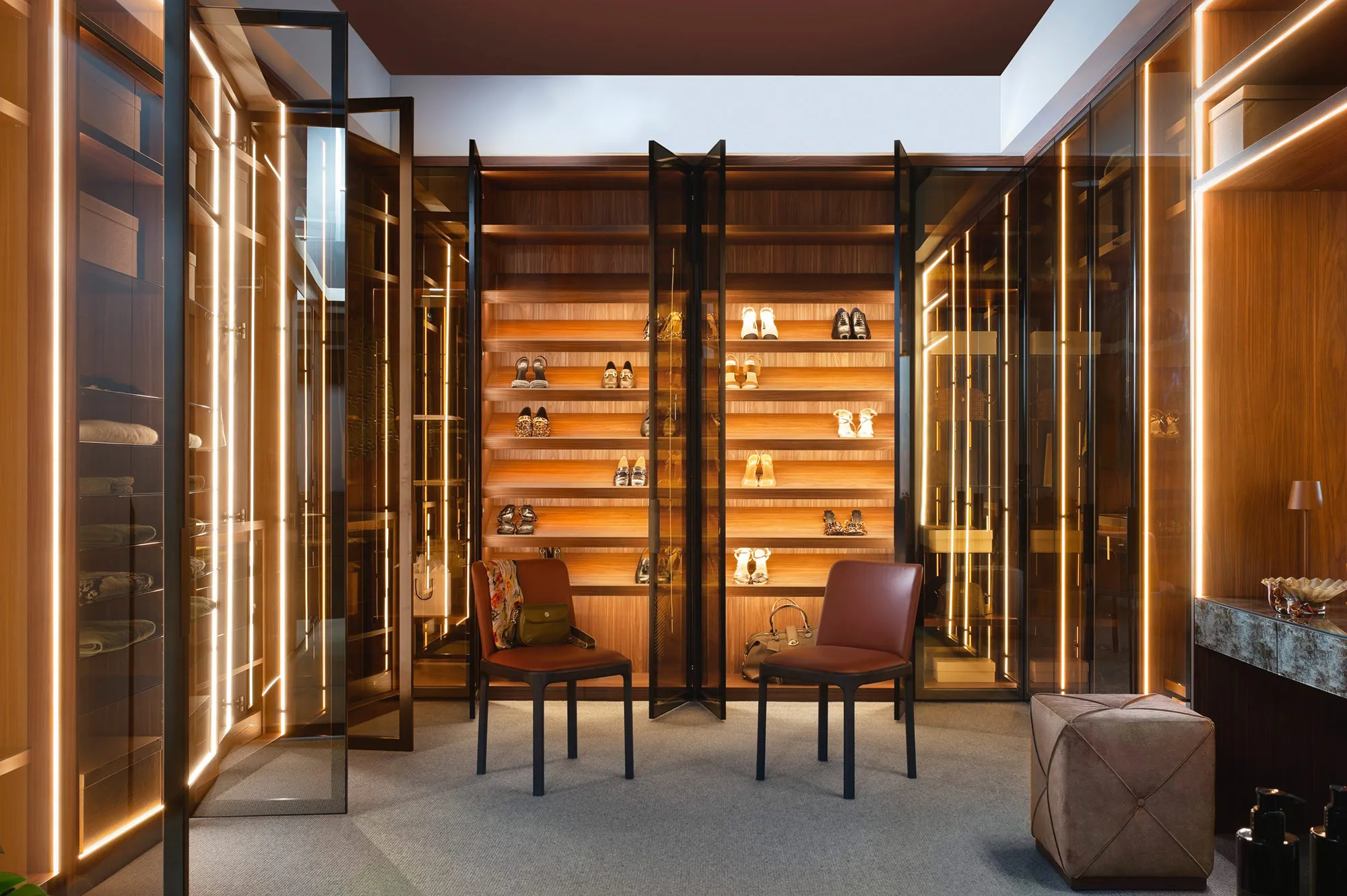 Walk-in dressing room with glass-fronted shelving, warm LED lighting, shoe displays and two leather chairs