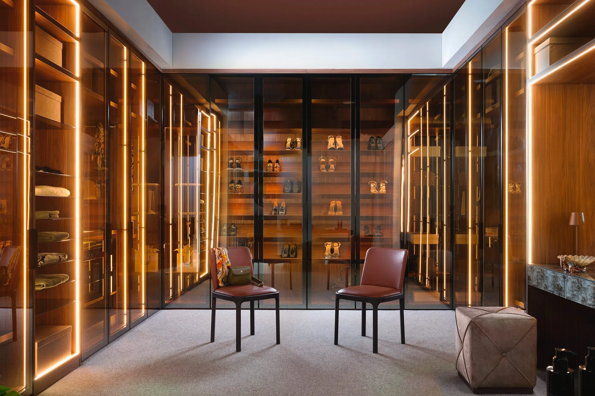 Walk-in wardrobe with smoked glass doors, warm LED lighting and two red leather chairs