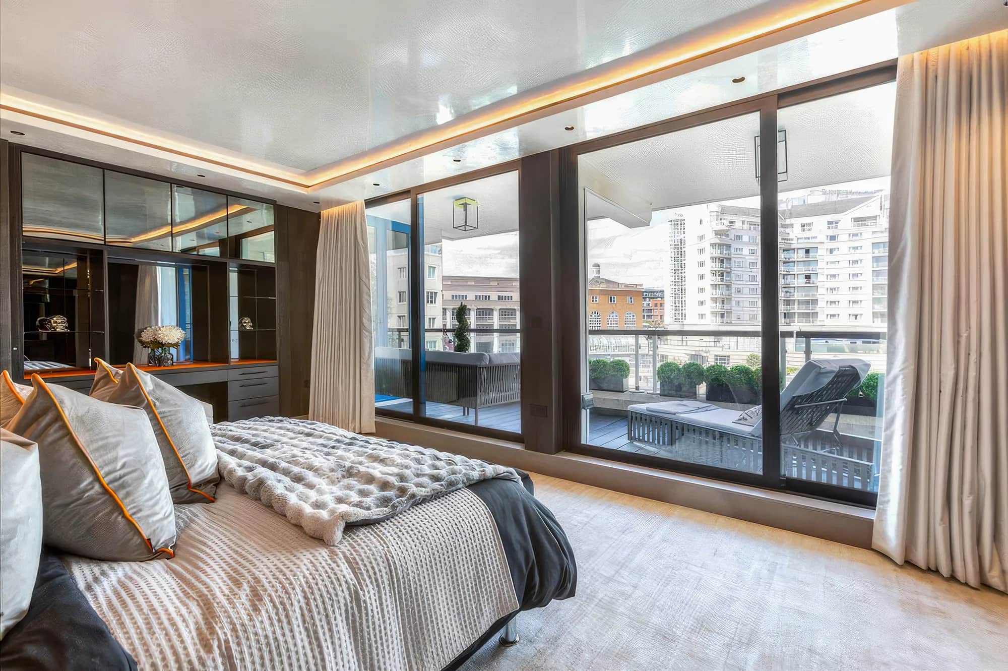 Penthouse bedroom with fitted wardrobes beside a balcony window overlooking riverside apartments