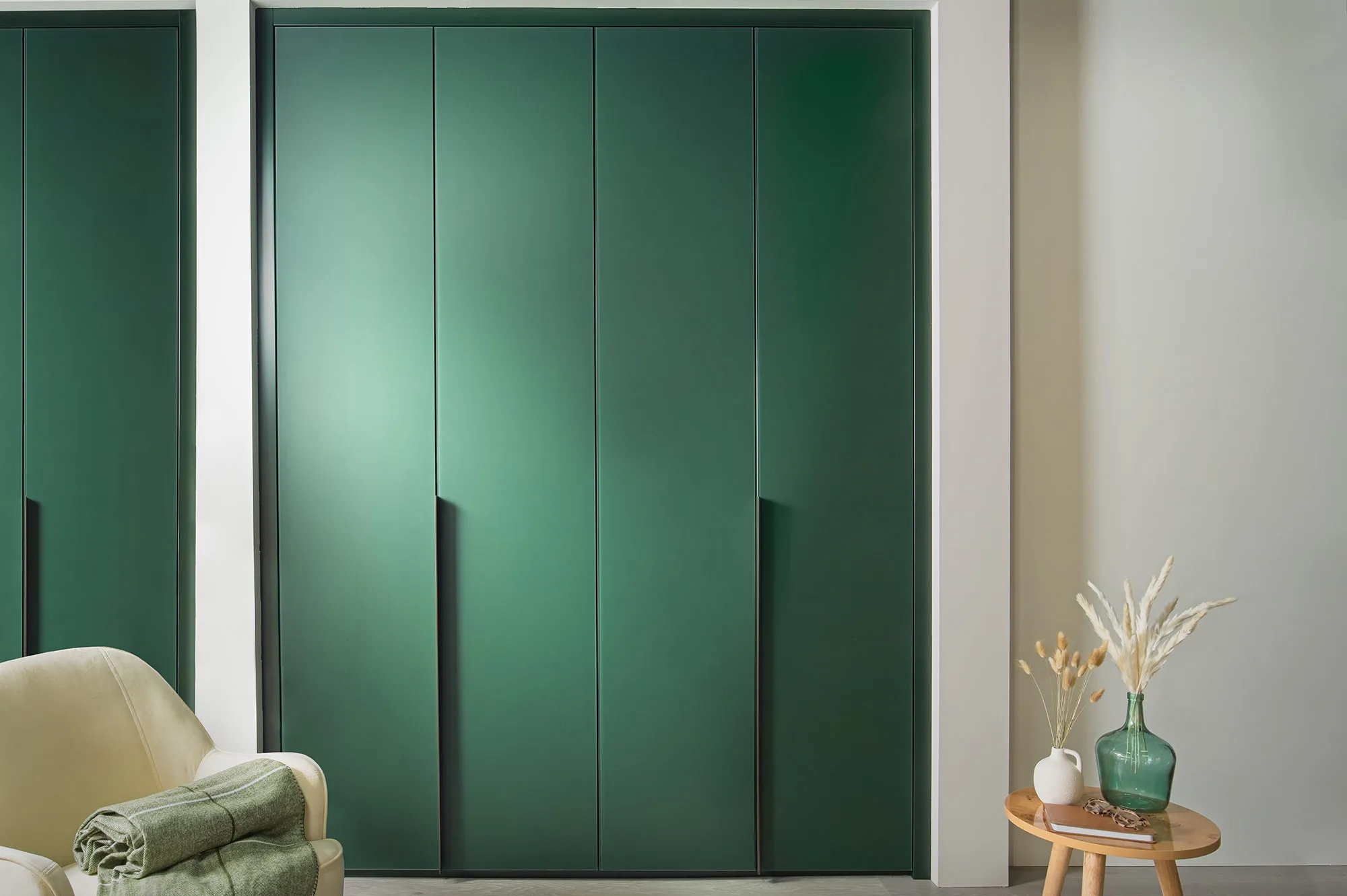 Floor-to-ceiling green fitted wardrobe doors with slim vertical handles beside a cream armchair